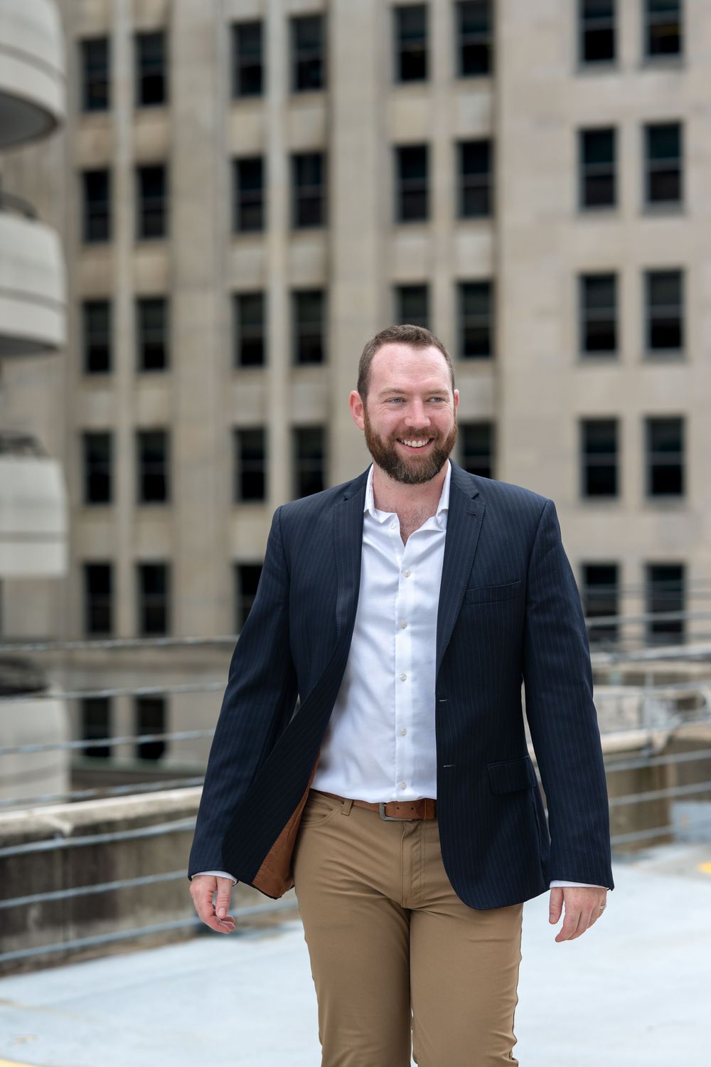 Smiling bearded man in navy blazer and khaki pants walking outdoors in front of a large urban building.