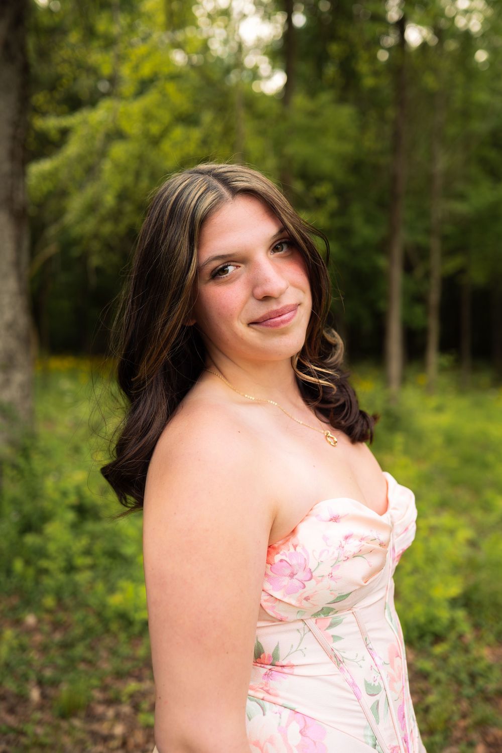 Young woman in floral strapless dress posing outdoors among lush green trees, smiling softly over her shoulder.