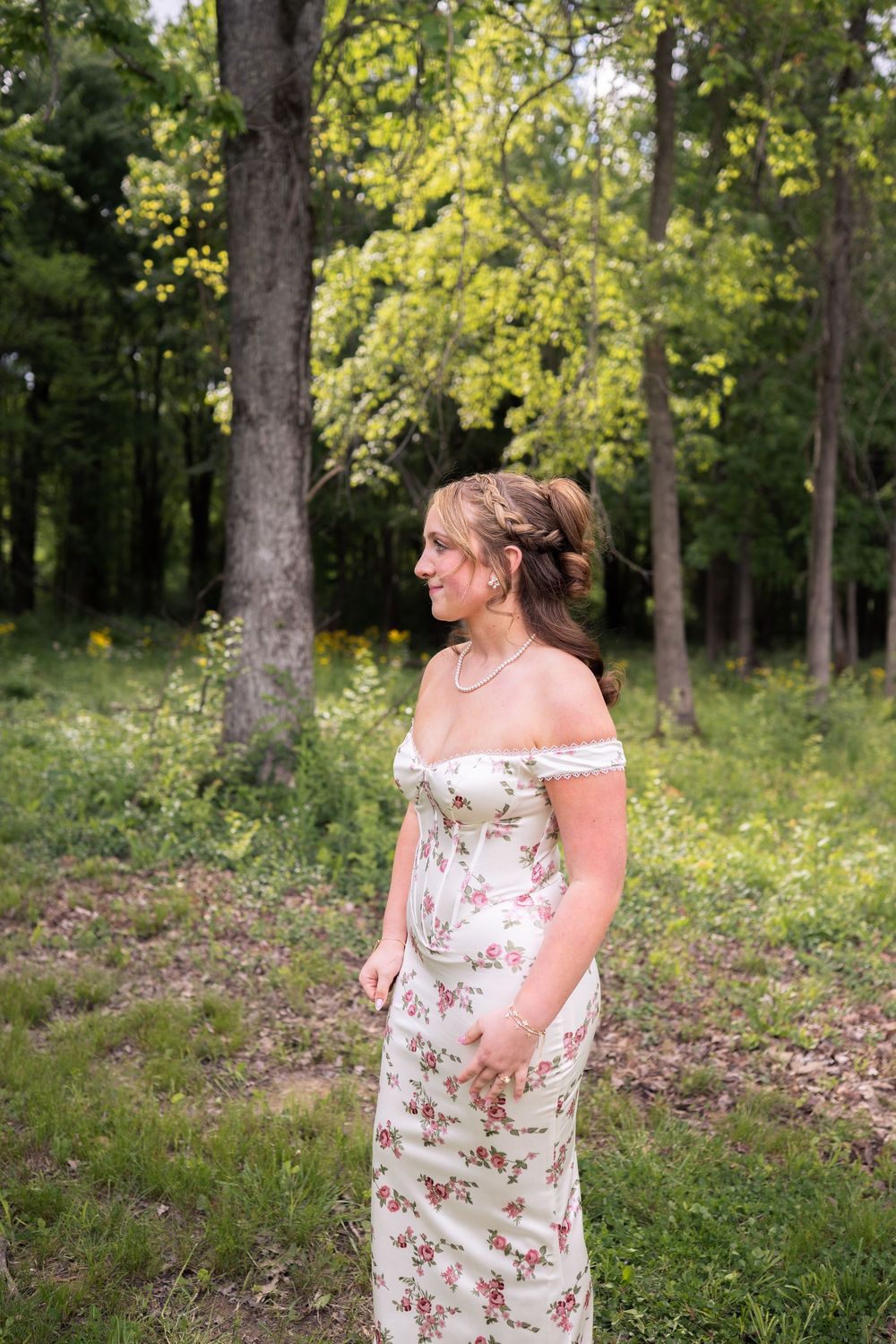 Young woman in floral off-shoulder maxi dress standing in a lush green forest clearing, looking to the side.