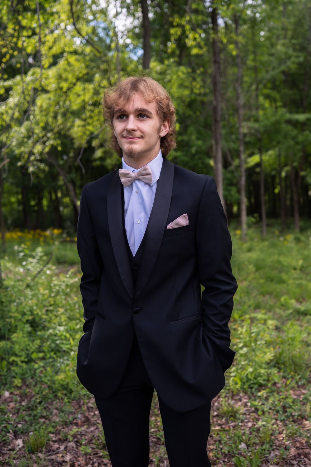 Young man in black tuxedo with bow tie and pink pocket square posing outdoors in a lush green forest setting.