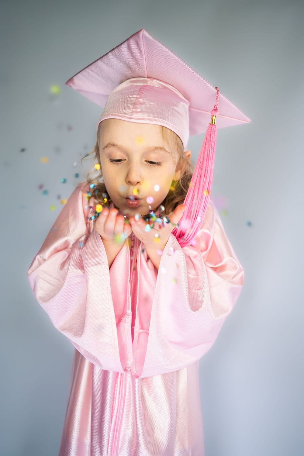 Young girl in pink graduation cap and gown blowing colorful confetti from her hands.