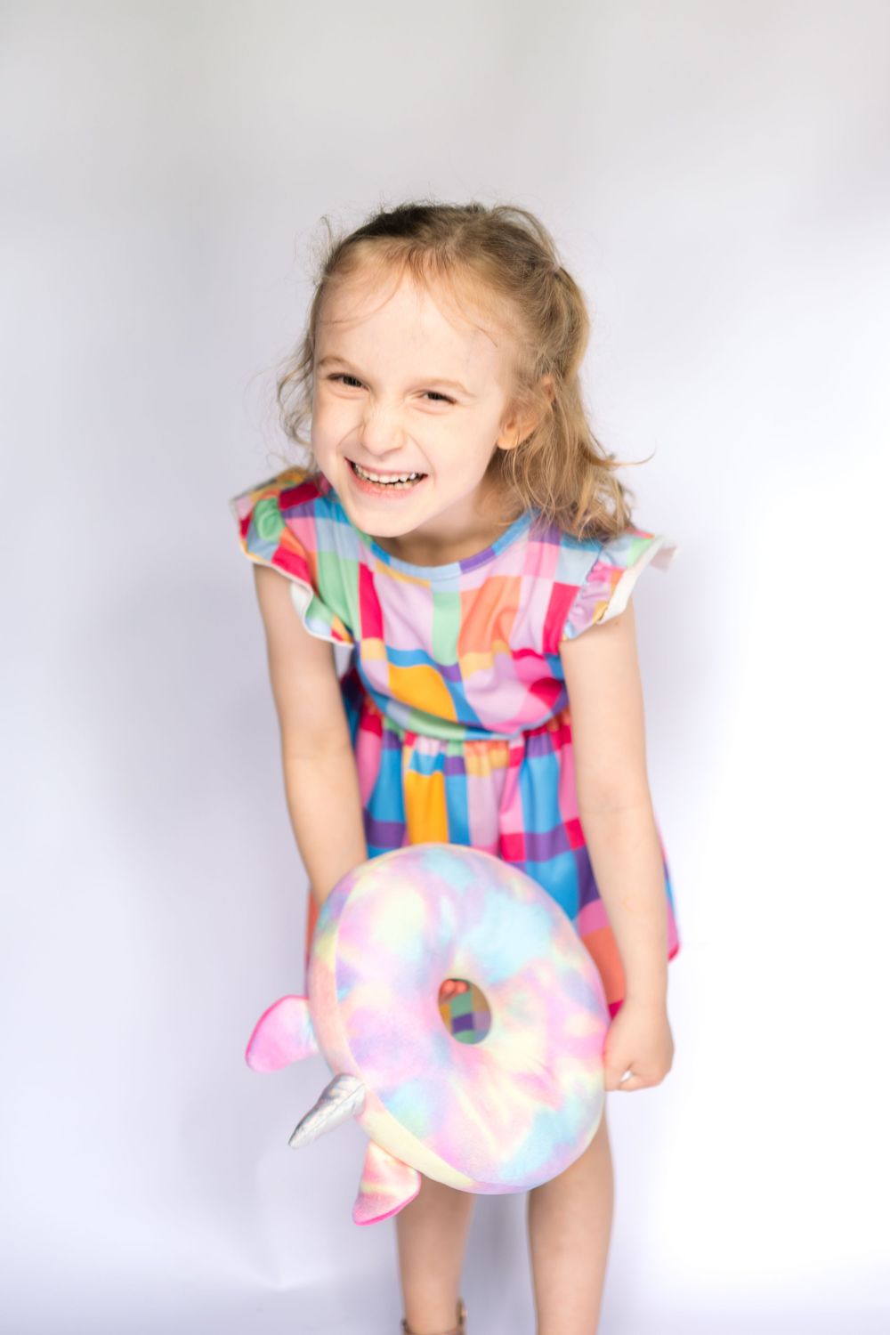 Happy young girl in colorful dress laughing while holding a pastel donut plush toy against white background.
