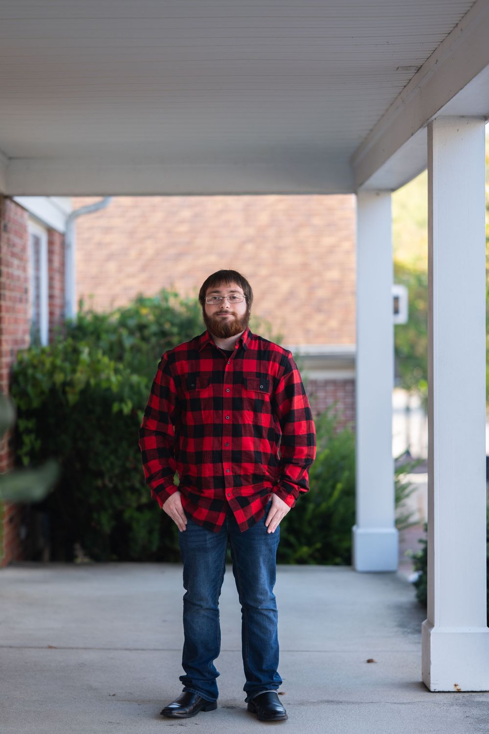 Man in red plaid flannel shirt and jeans standing on a covered porch with brick building and greenery behind him.