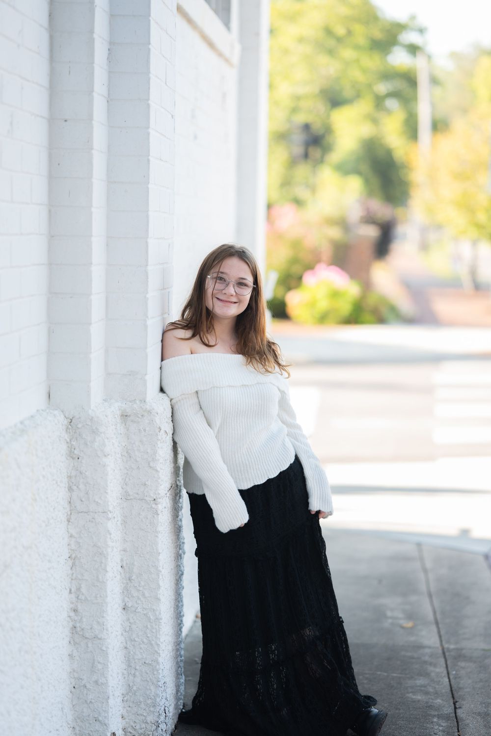 Young woman in white off-shoulder sweater and black maxi skirt leaning against a white brick wall outdoors.