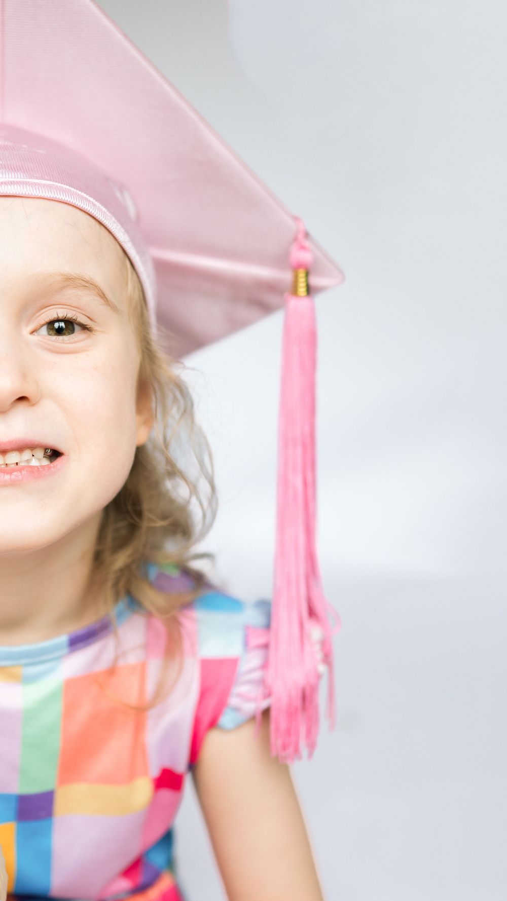 A young girl smiling in a pink graduation cap and colorful dress, celebrating her kindergarten graduation.