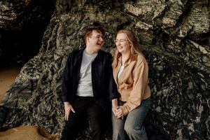 Smiling couple holding hands while sitting against dramatic dark rock face on beach.