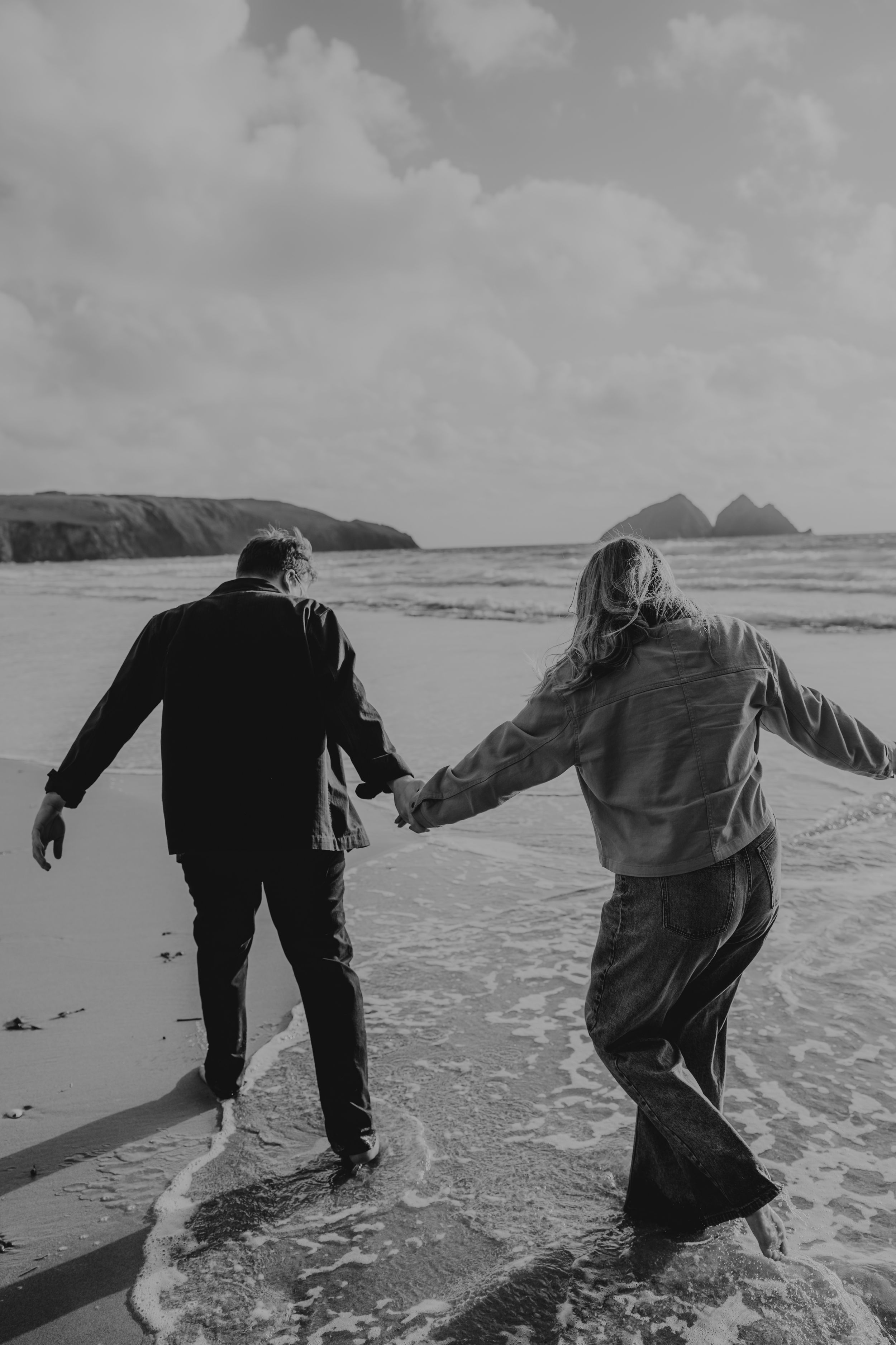 Black and white photo of couple holding hands walking in ocean waves on a rocky beach.