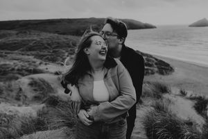 Black and white photo of a couple sharing a romantic moment on a scenic beach with rolling hills in the background.