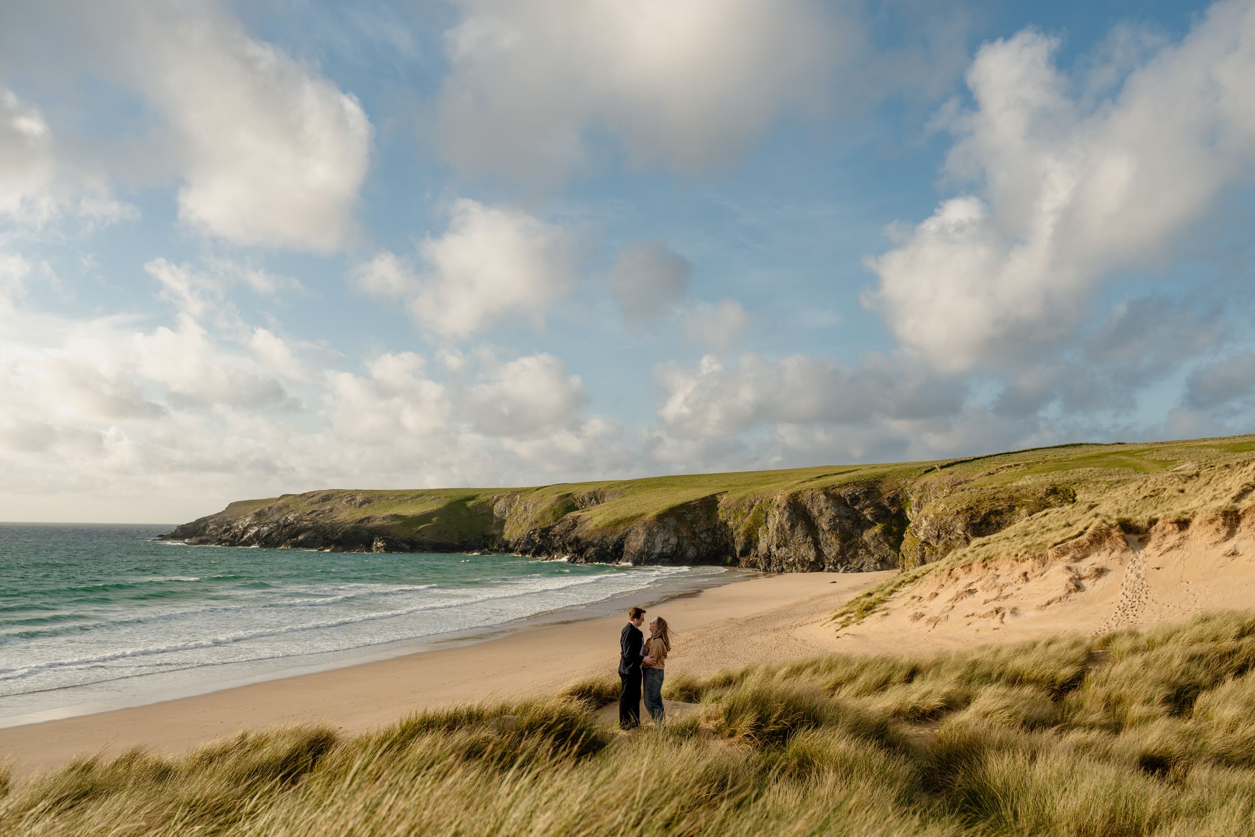 Couple standing together on grassy dunes overlooking a stunning curved sandy beach and headland.