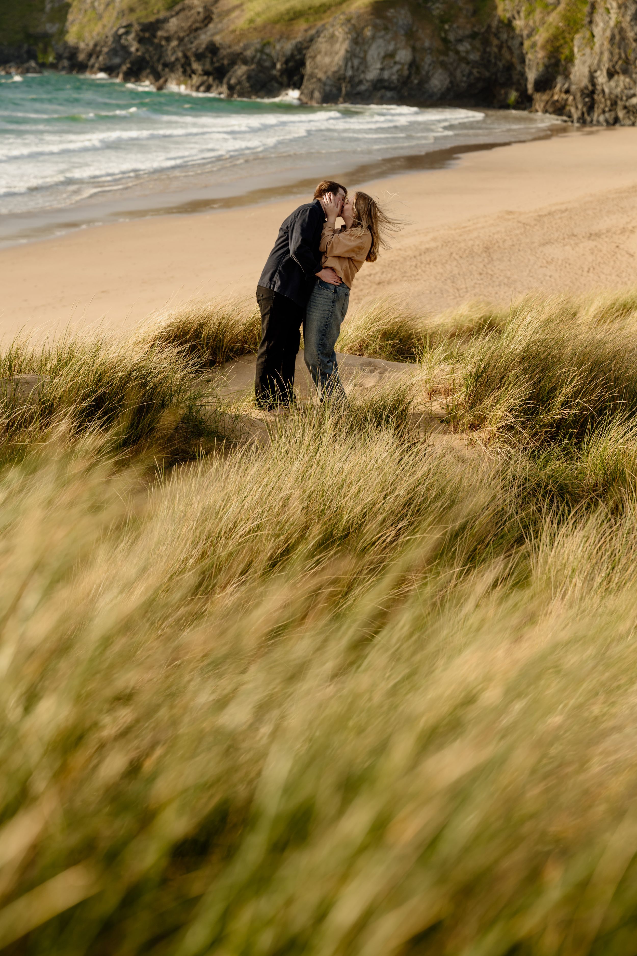 Romantic couple embracing in golden grass dunes with scenic sandy beach and ocean behind them.