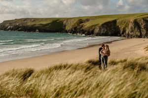 Couple walking through coastal dunes with a scenic sandy beach and green cliffs beyond.