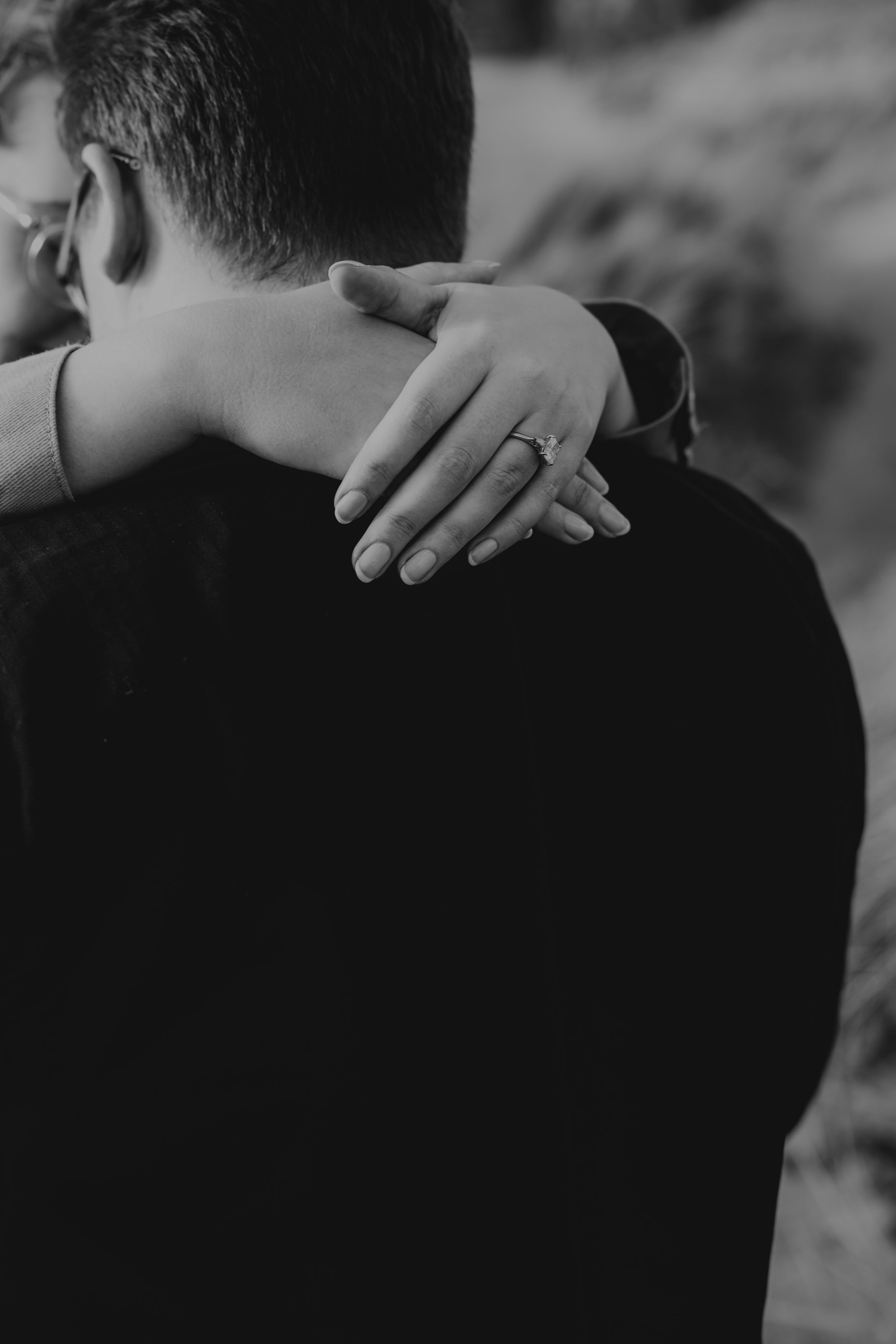 Black and white photo of woman embracing man from behind showing engagement ring on hand.