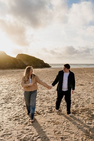 Couple holding hands and smiling at each other while walking barefoot on a sunny sandy beach.