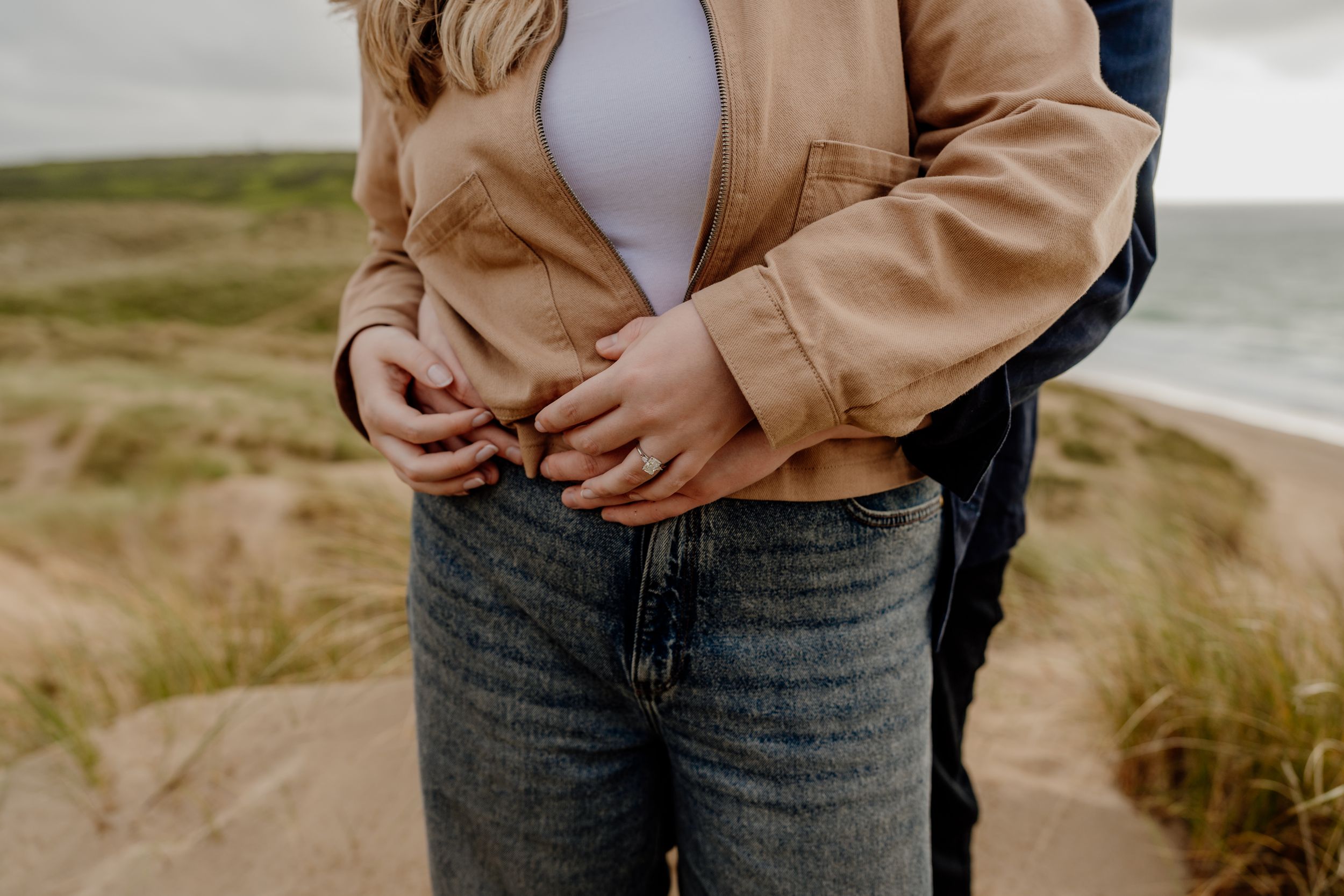 Couple embracing on a sandy beach, woman in tan jacket and jeans with partner's arms wrapped around her waist.