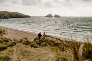 Two people walk along a scenic sandy beach with dramatic rocky islands visible in the calm ocean under overcast skies.