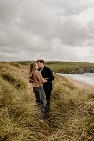 Couple kissing in coastal grass dunes with sandy beach and dramatic cliffs in the background.