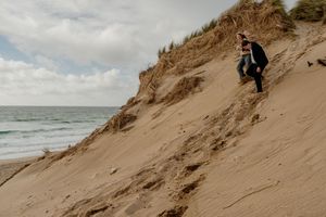 Couple climbing a sandy coastal dune with ocean waves in the background on a cloudy day.