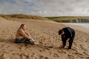 Woman sitting on beach sand while man bends down beside her with green cliffs in background.