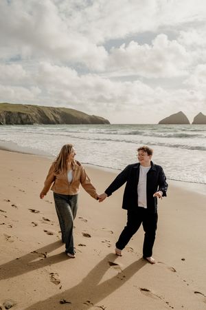 Couple holding hands and walking barefoot on a sandy beach with rocky cliffs and ocean waves in the background.