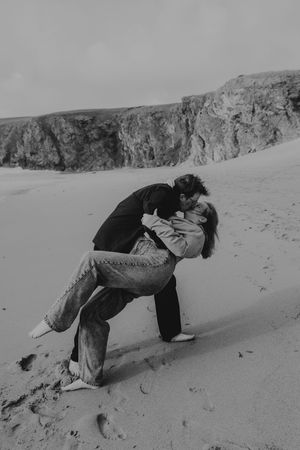 Black and white photo of couple sharing a romantic dip kiss on sandy beach with dramatic cliffs behind.