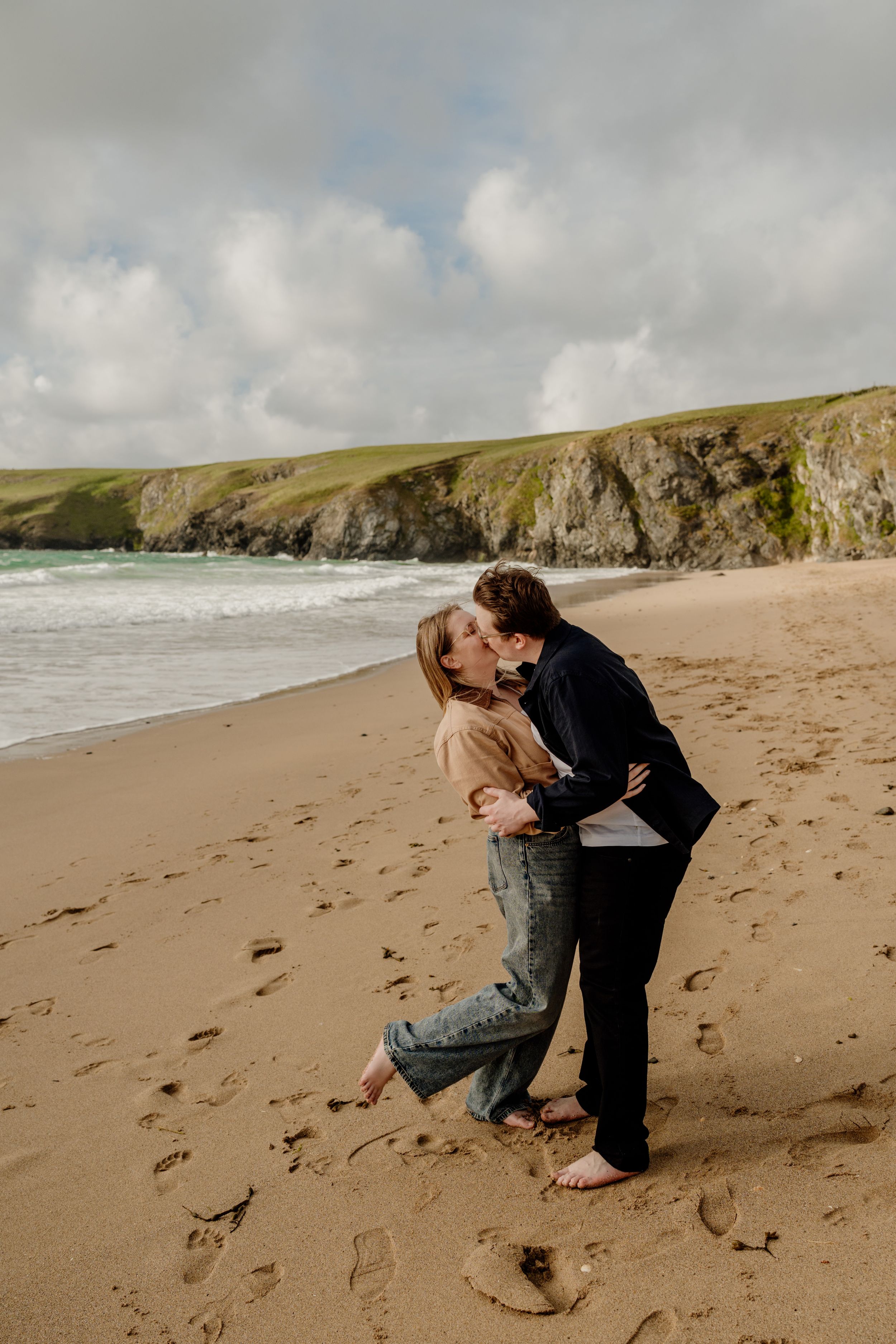 Romantic couple sharing a kiss on a sandy beach with dramatic cliffs and crashing waves behind them.