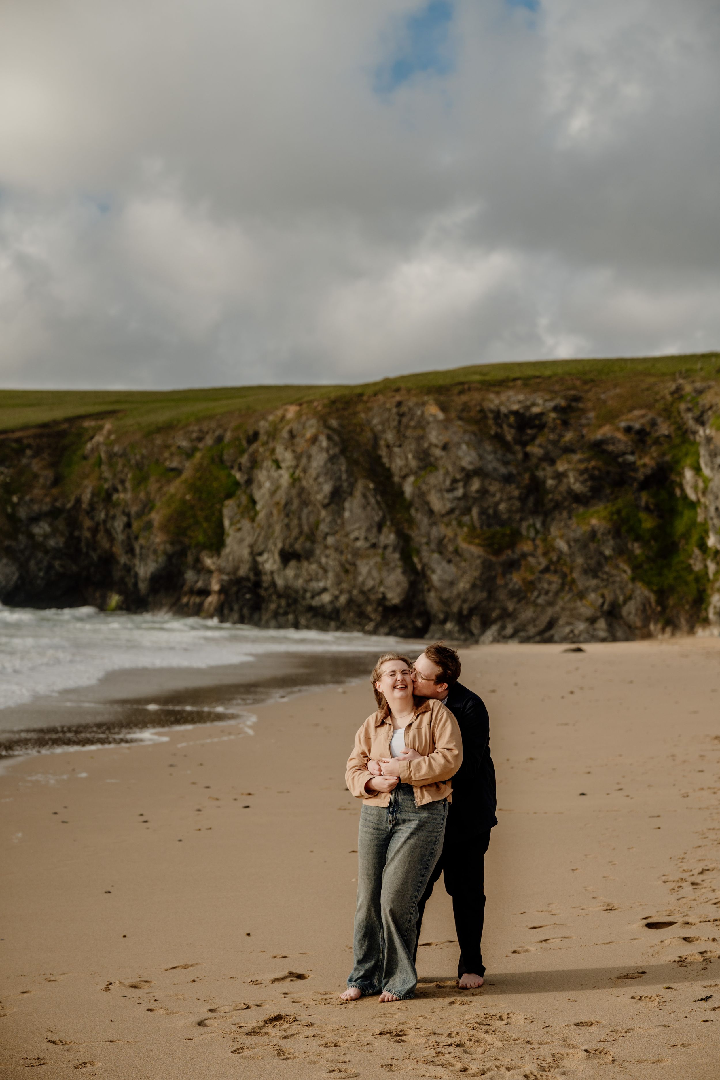 Couple embracing on a sandy beach with dramatic cliffs and ocean waves in the background.