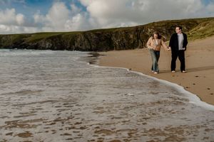 Couple strolling hand in hand along a sandy beach with dramatic rocky cliffs and crashing waves in background.