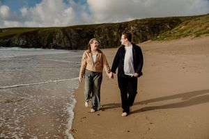 Happy couple holding hands walking along shoreline with dramatic green cliffs behind them.