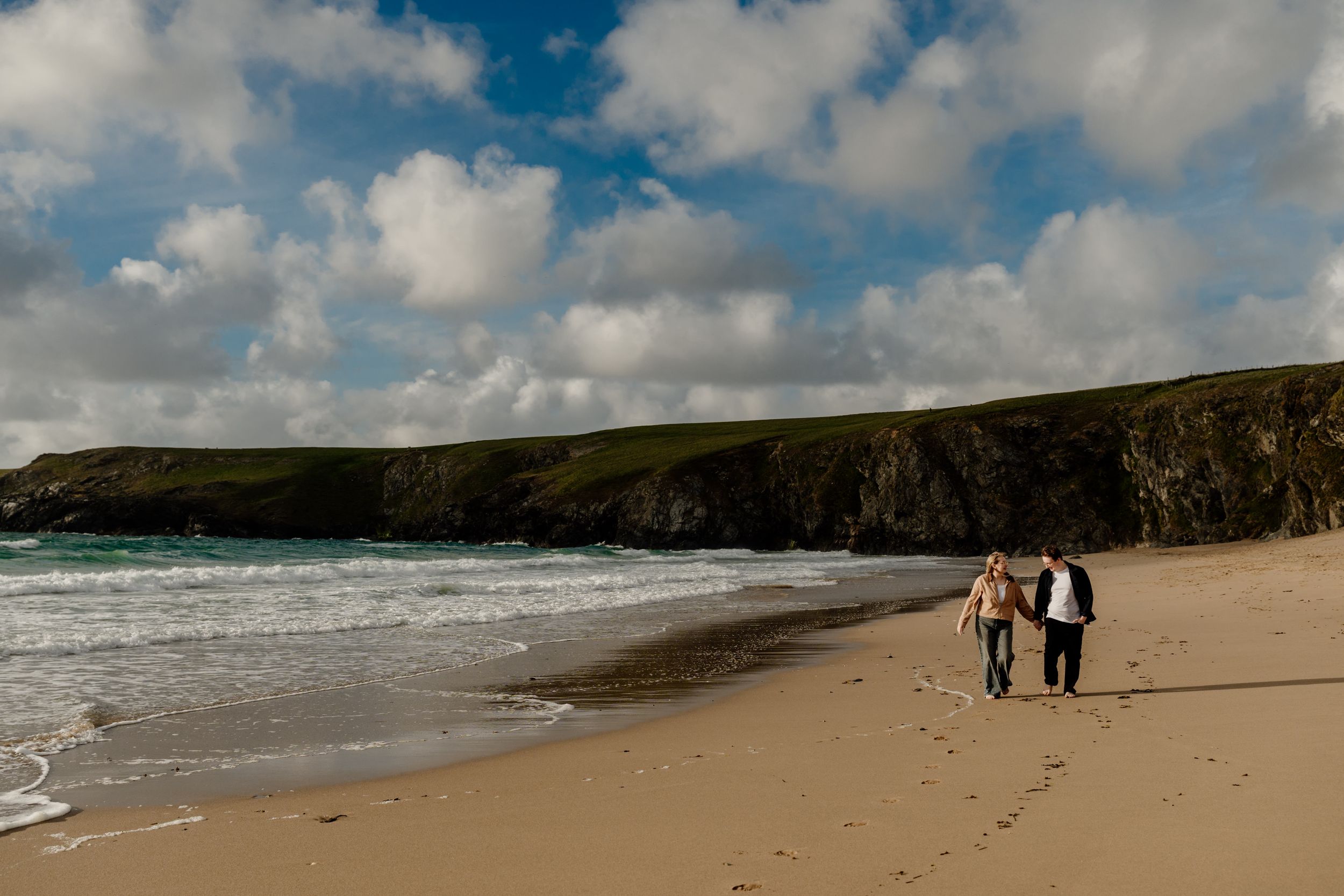 Couple walking together on a wide sandy beach with stunning green cliffs and dramatic cloudy blue sky backdrop.