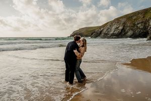 Couple kissing on a sandy beach with ocean waves and dramatic cliffs in the background.