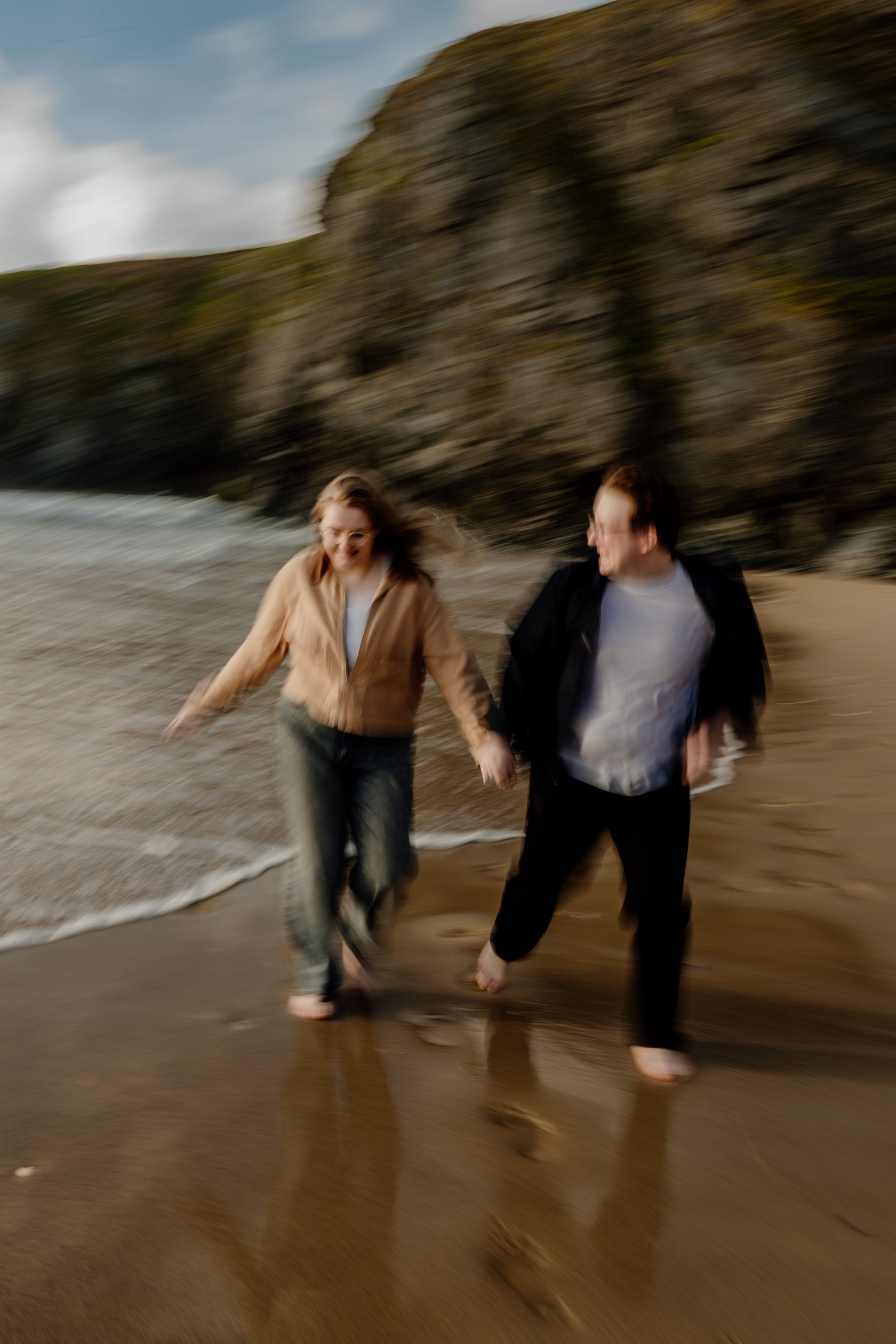 Couple running barefoot on wet sandy beach with rocky cliffs, captured with motion blur effect.