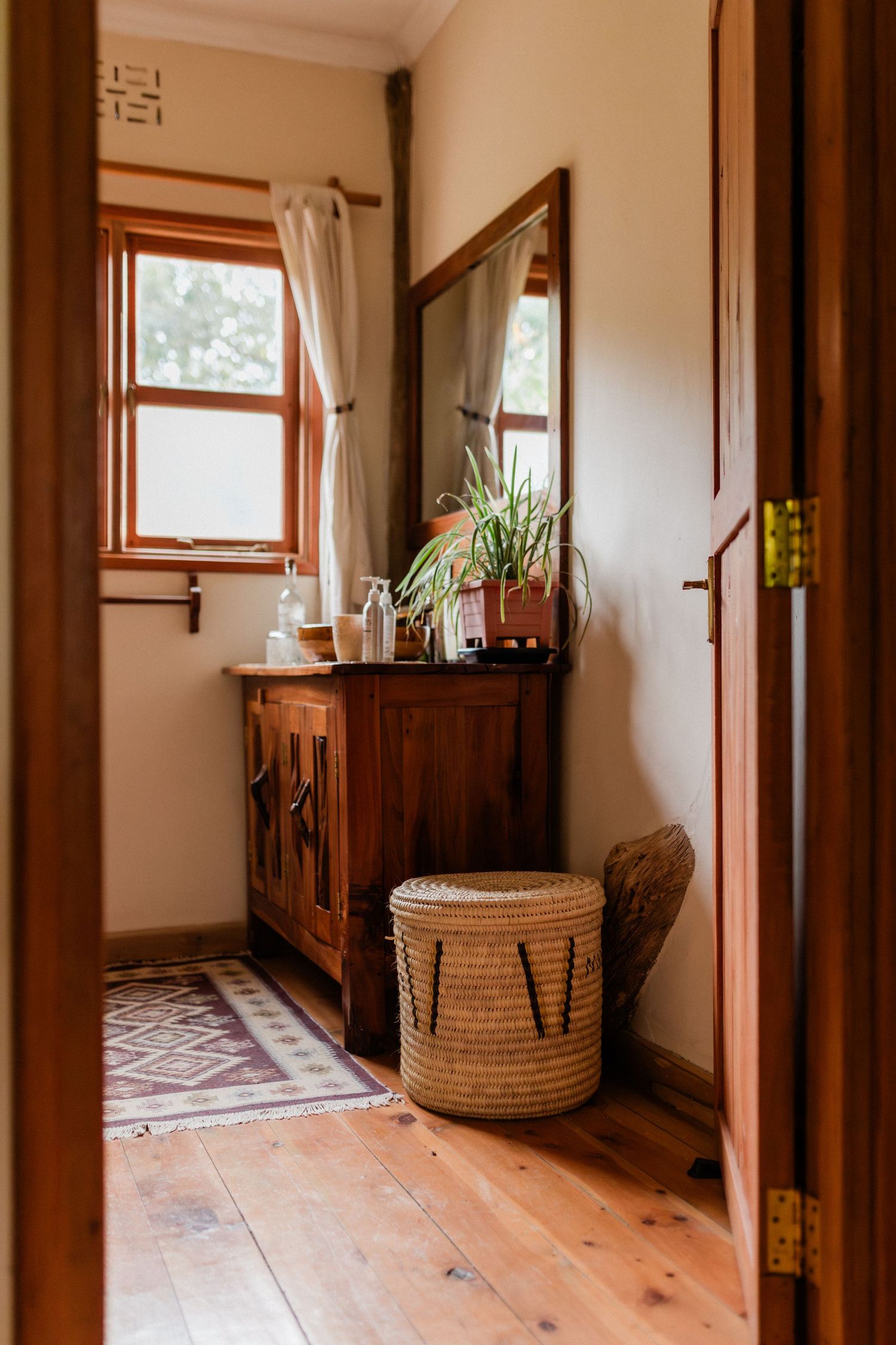 Warm bohemian bathroom with wooden cabinet, wicker basket, hardwood floors, mirror, and plant near a sunlit window.