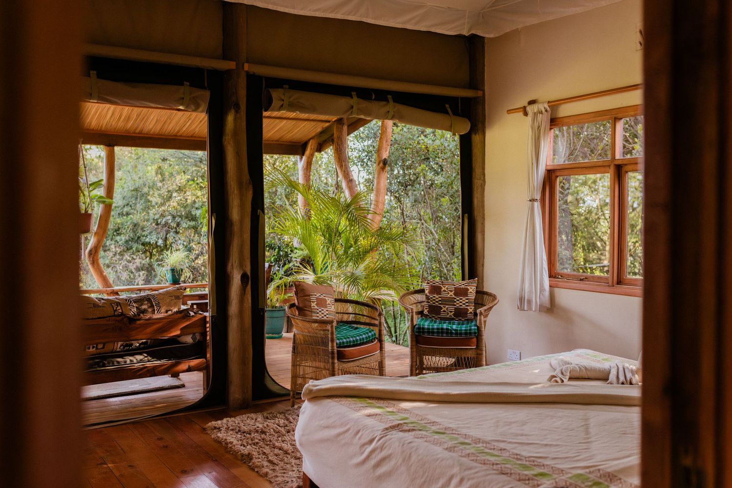 Cozy jungle bedroom with open doors, wicker chairs, lush greenery views, wooden floors, and a white bed with natural light.
