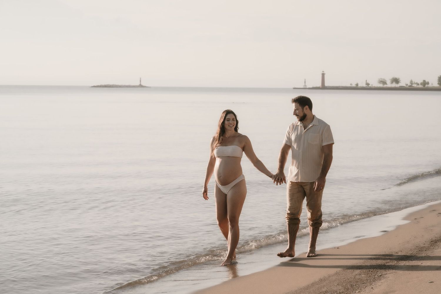 Couple holding hands walking along sandy beach shoreline at sunset, woman in white bikini, man in casual shirt.