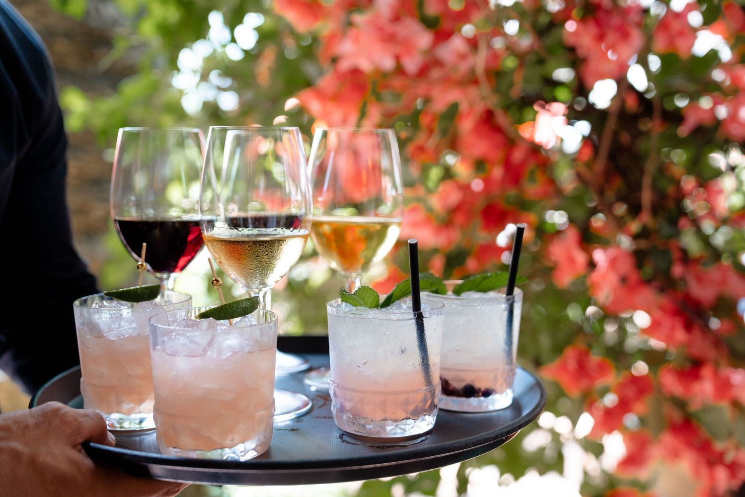A server holds a tray of cocktails and wine glasses outdoors with colorful red flowers in the background.