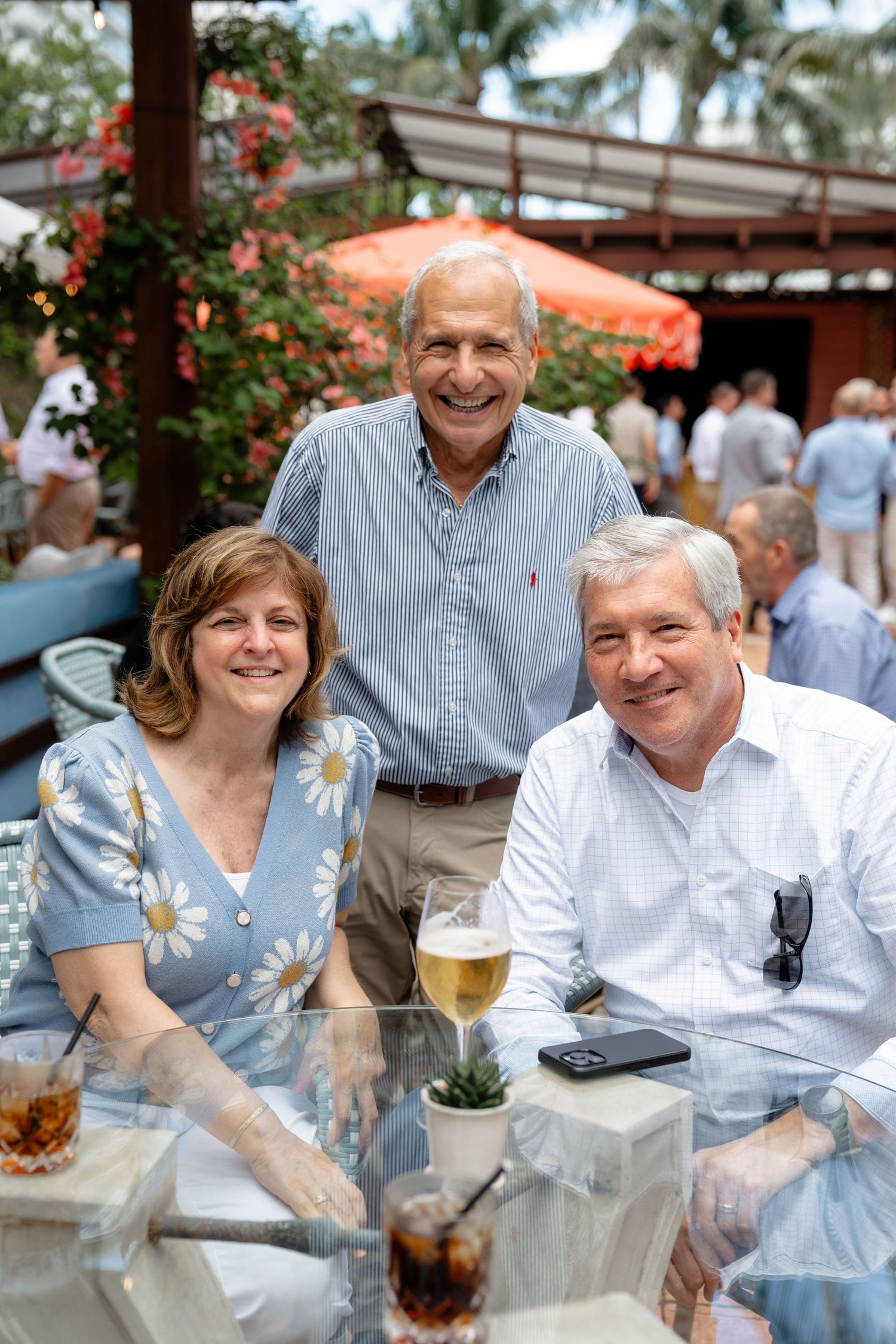 Three smiling adults enjoying drinks at an outdoor restaurant patio with tropical greenery.