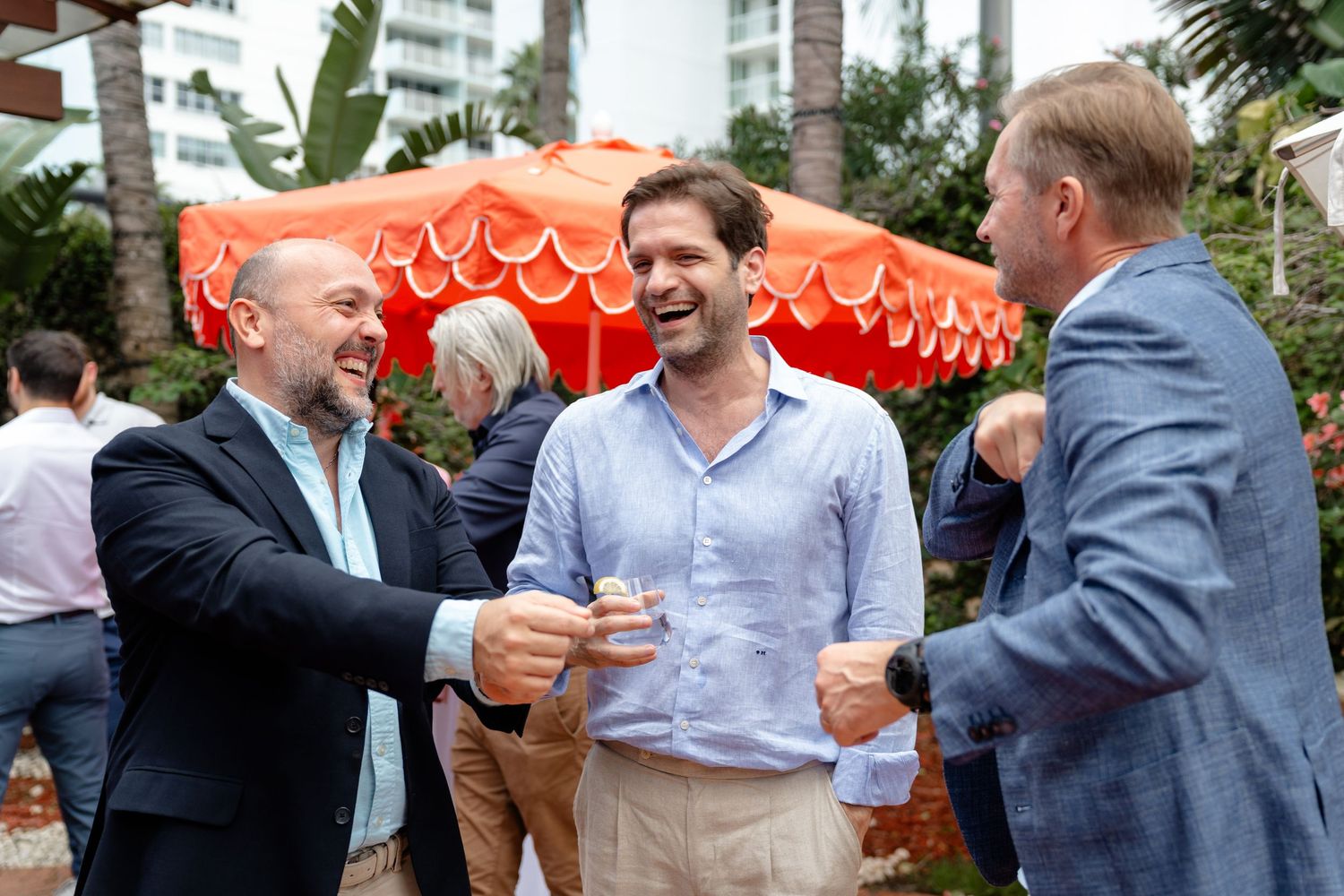 Three men laughing and toasting drinks at an outdoor social event with colorful orange umbrella backdrop.