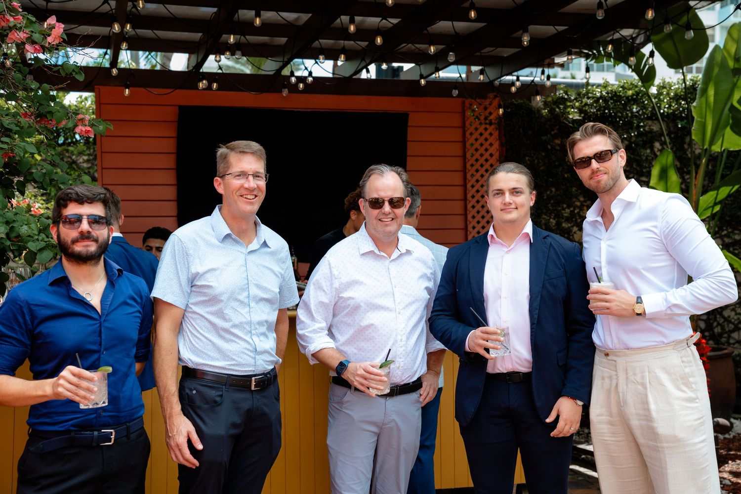Five men smiling and holding drinks at an outdoor corporate networking event under a pergola.