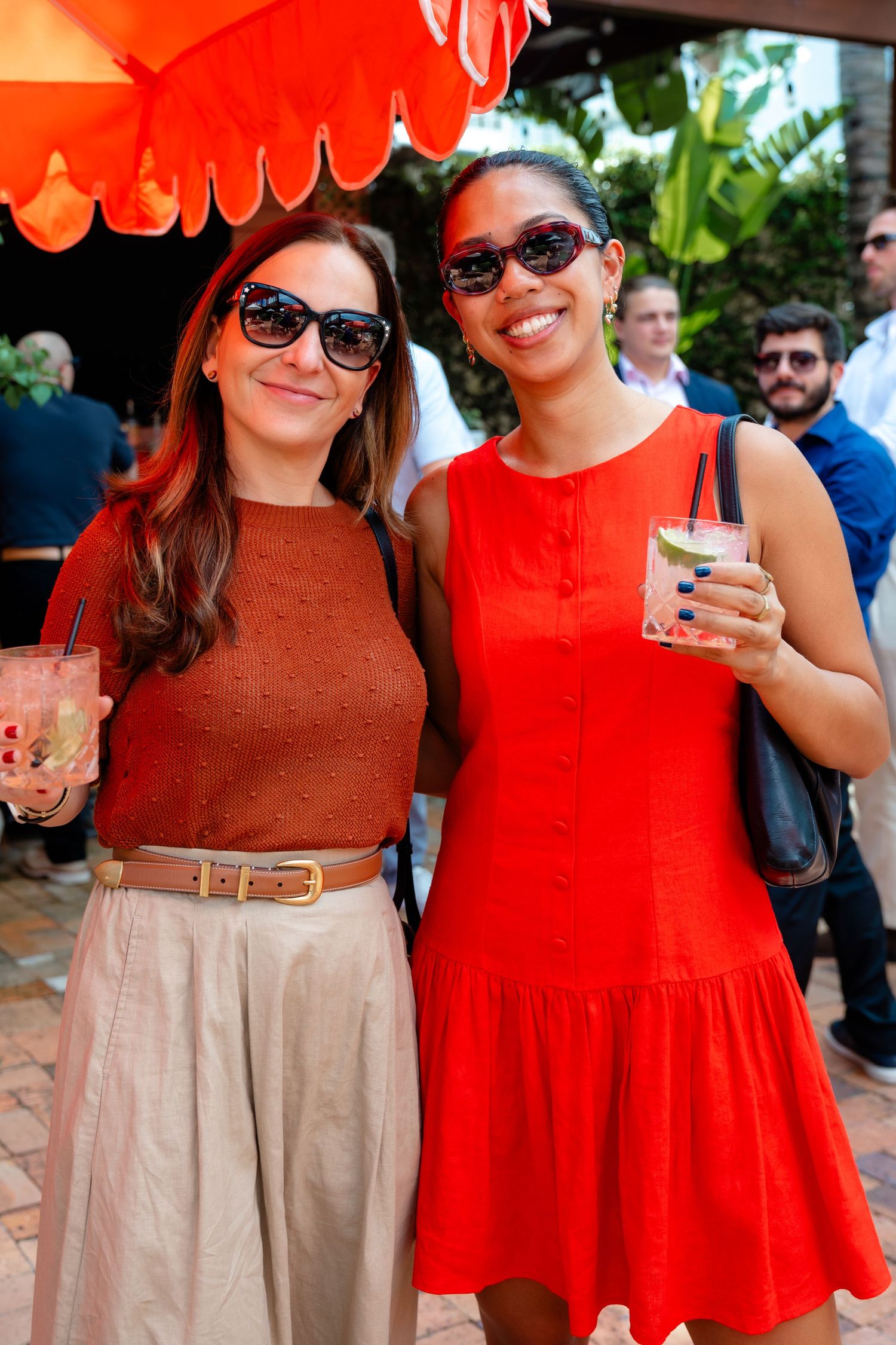 Two women in sunglasses smile holding cocktails at an outdoor summer party under an orange umbrella.