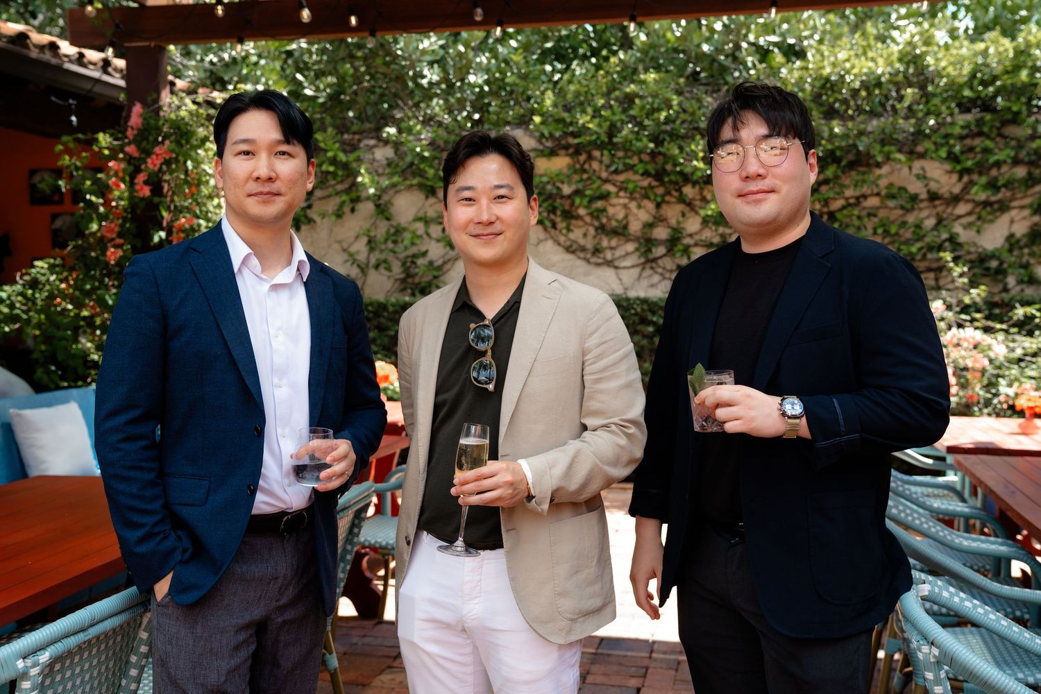 Three men smiling and holding drinks at an outdoor garden party event.