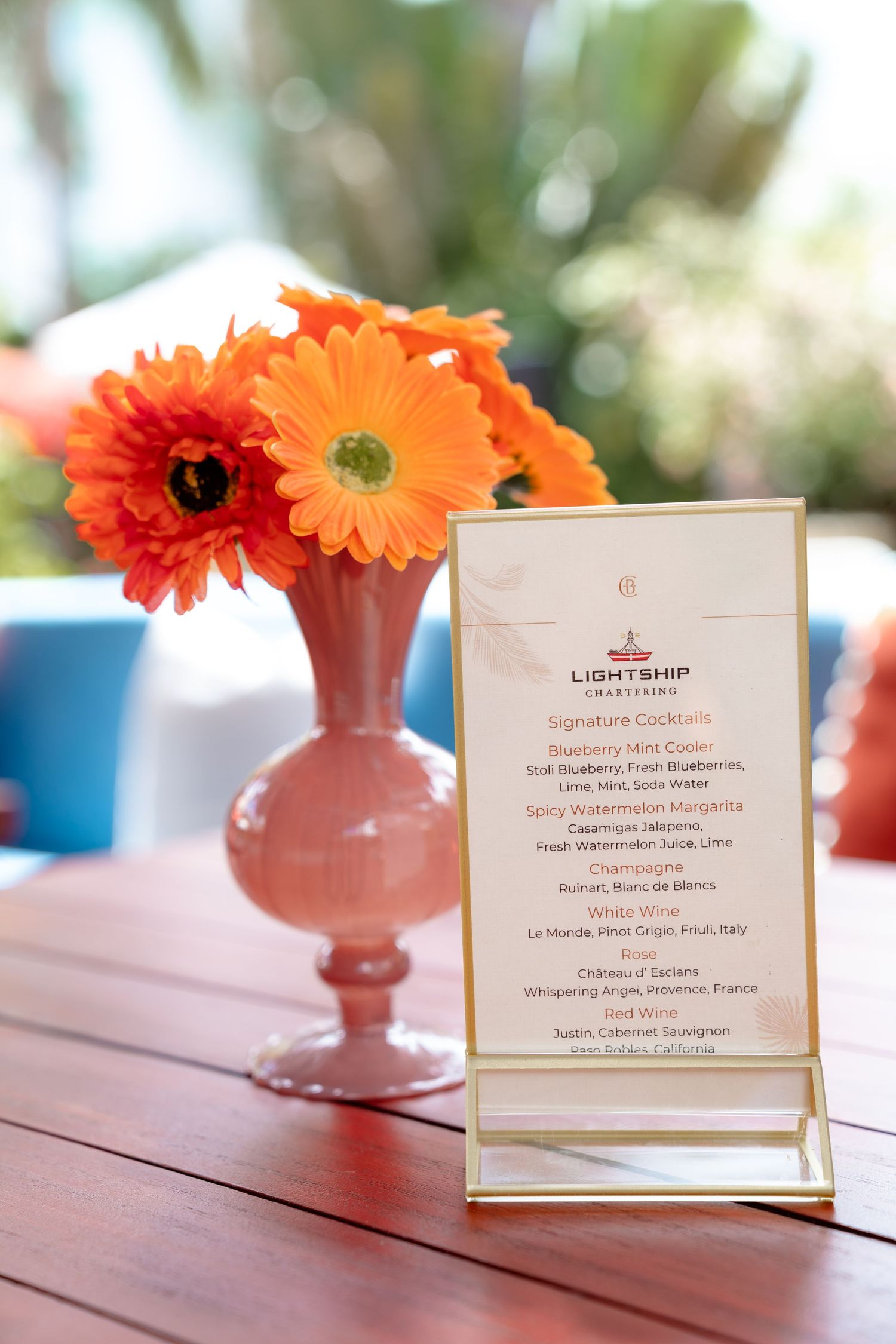 Orange gerbera daisies in pink vase beside Lightship Charters cocktail menu card on wooden table at outdoor event.
