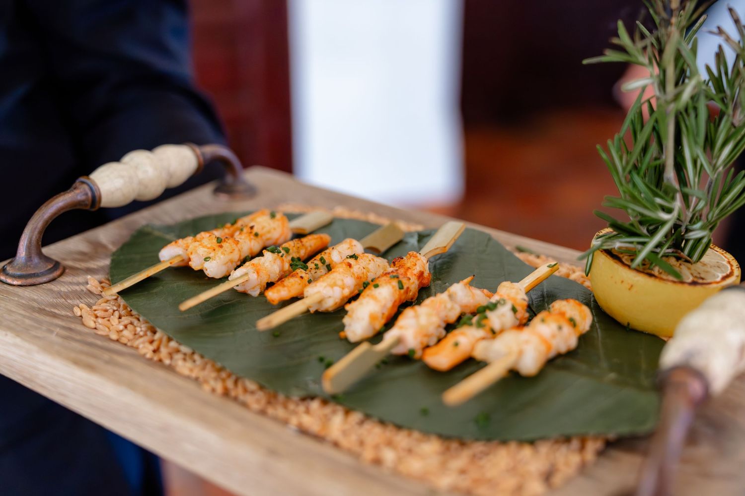 Grilled shrimp skewers on banana leaf with rosemary garnish served on a rustic wooden board.