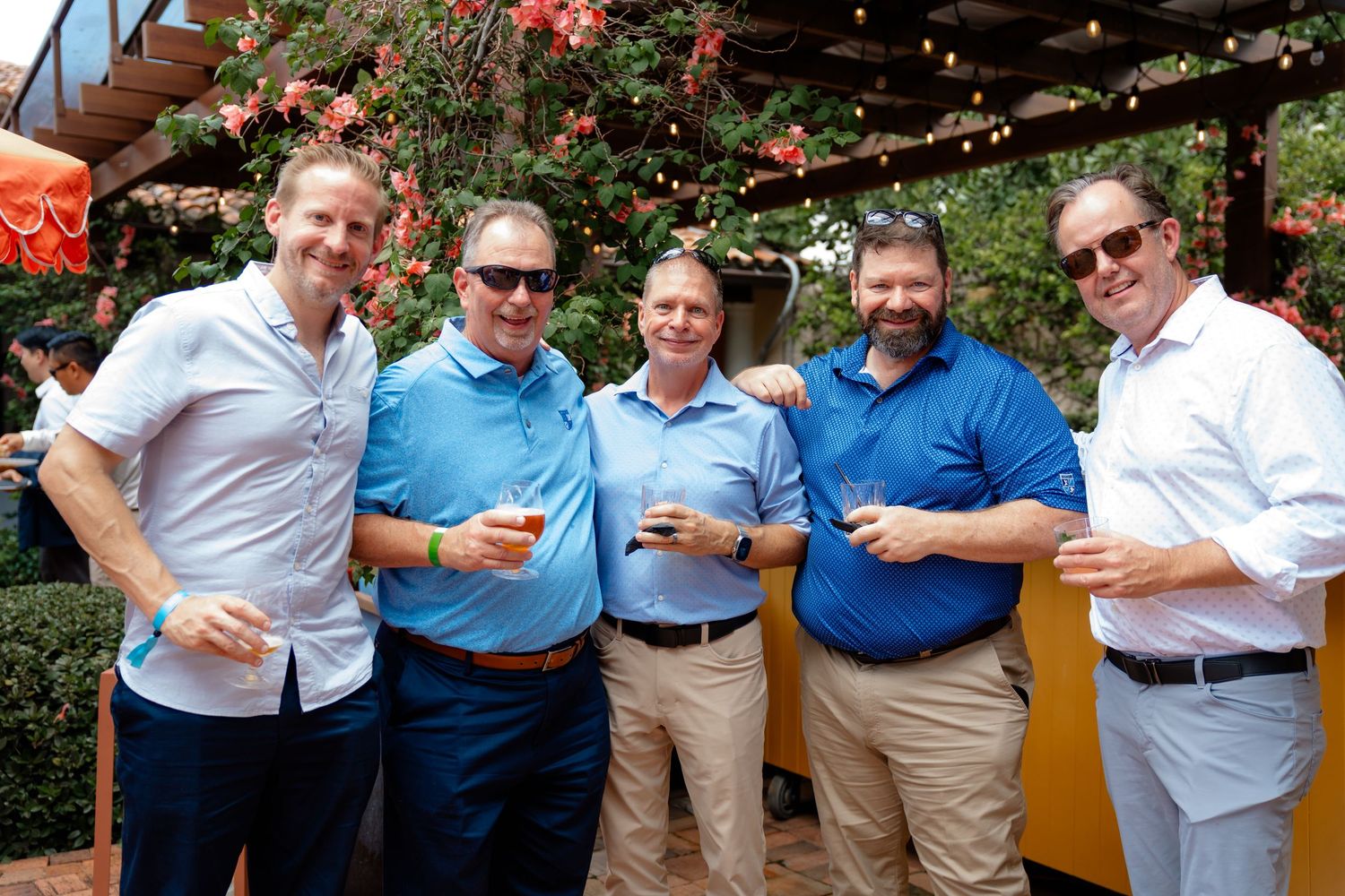 Five men smiling and holding drinks at an outdoor garden party event with floral decorations.