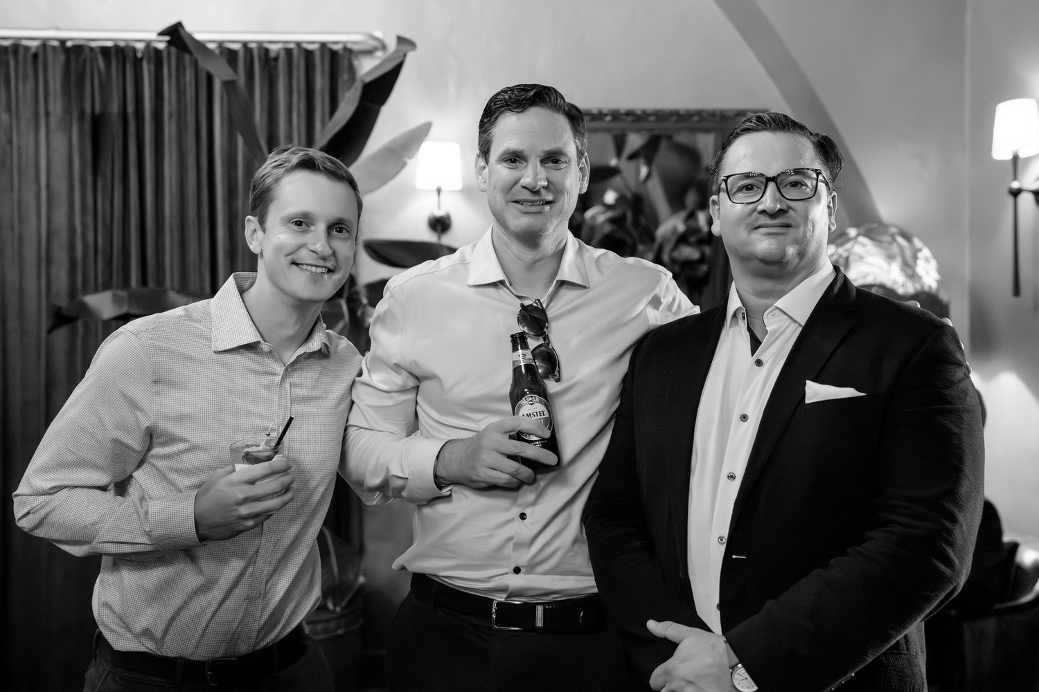 Three men smiling and posing together at a formal event, holding drinks in a black and white photo.