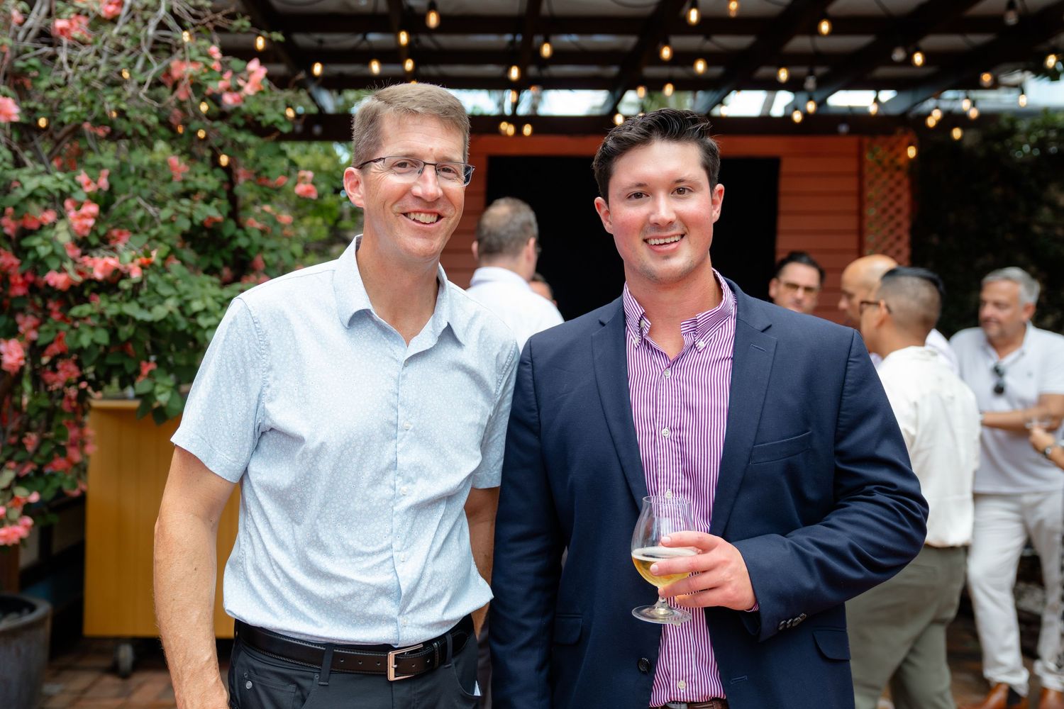 Two smiling men in smart casual attire pose together at an outdoor social gathering with string lights and flowers.