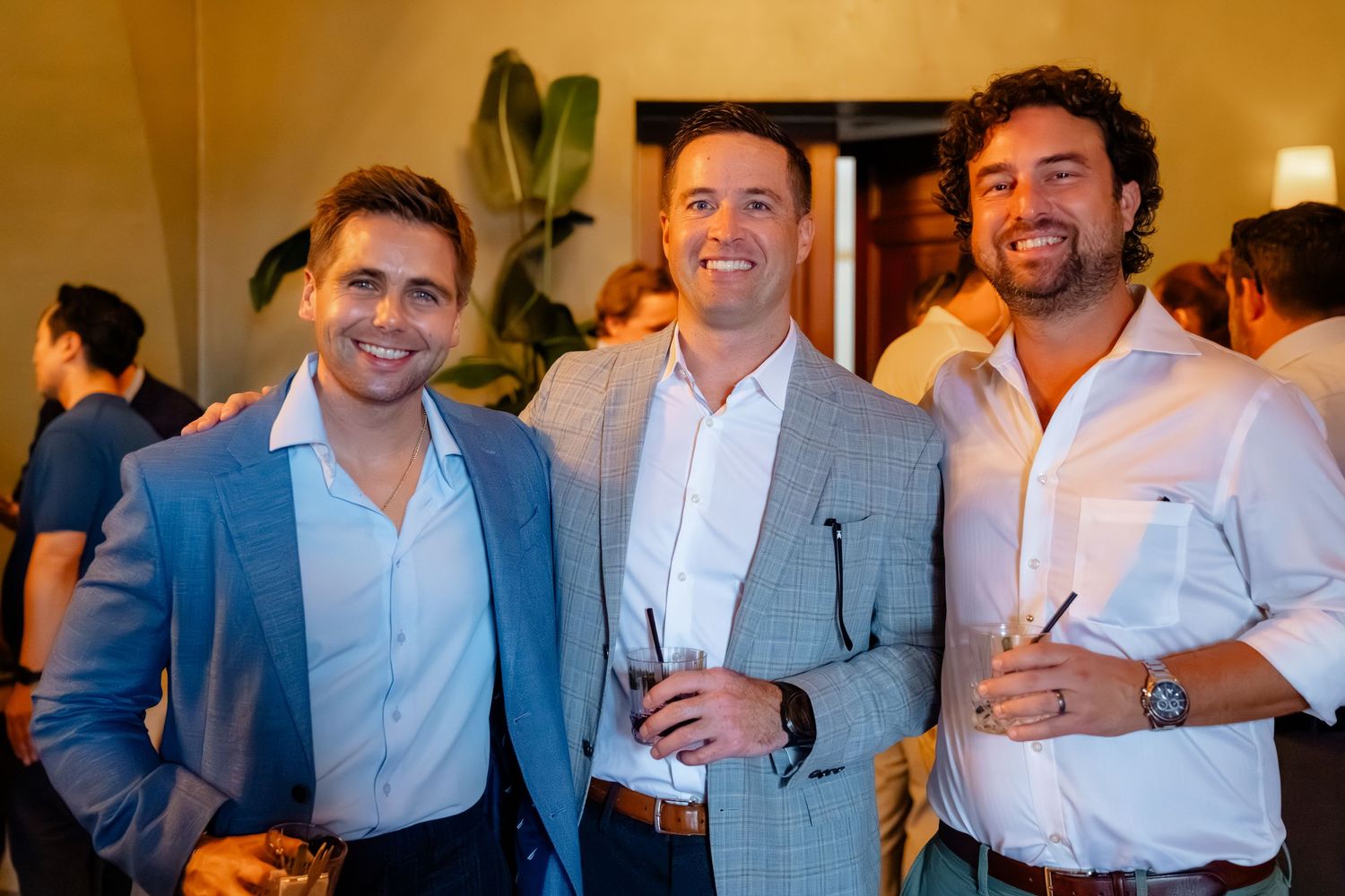 Three smiling men in blazers holding drinks at an indoor social gathering or networking event.