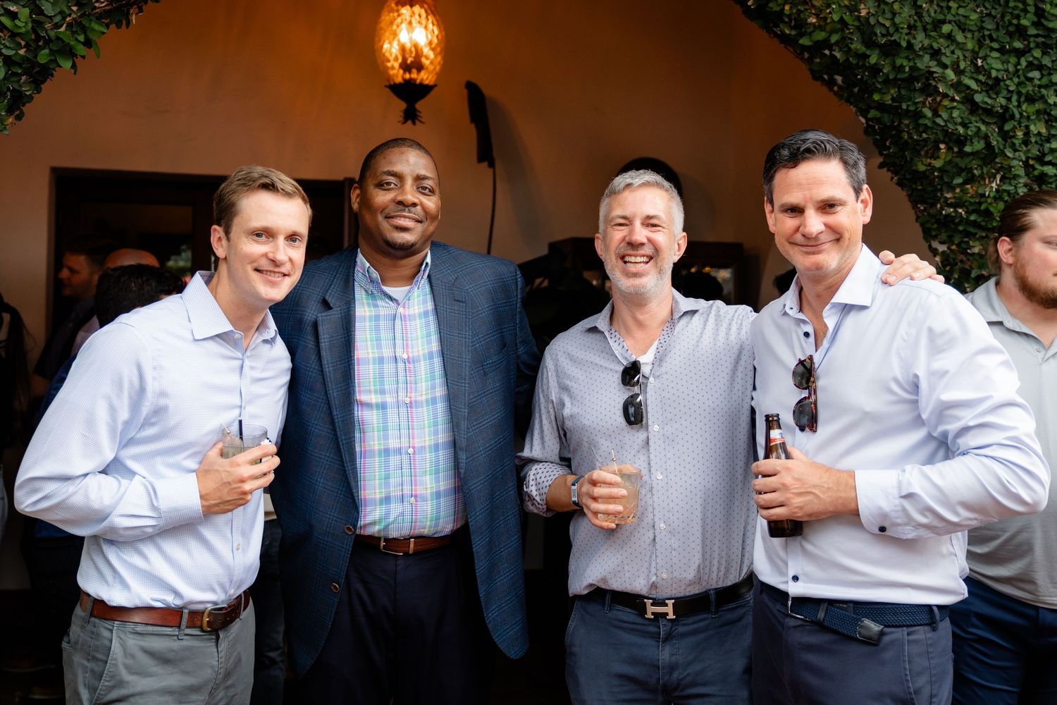 Four smiling men in business casual attire posing together outdoors at a social event, holding drinks.