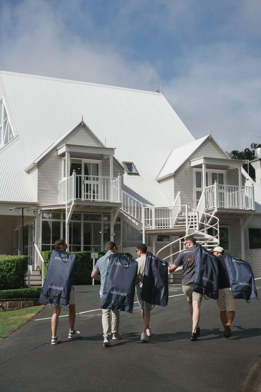 Groomsmen carrying garment bags walking toward a white colonial-style wedding venue on a cloudy day.