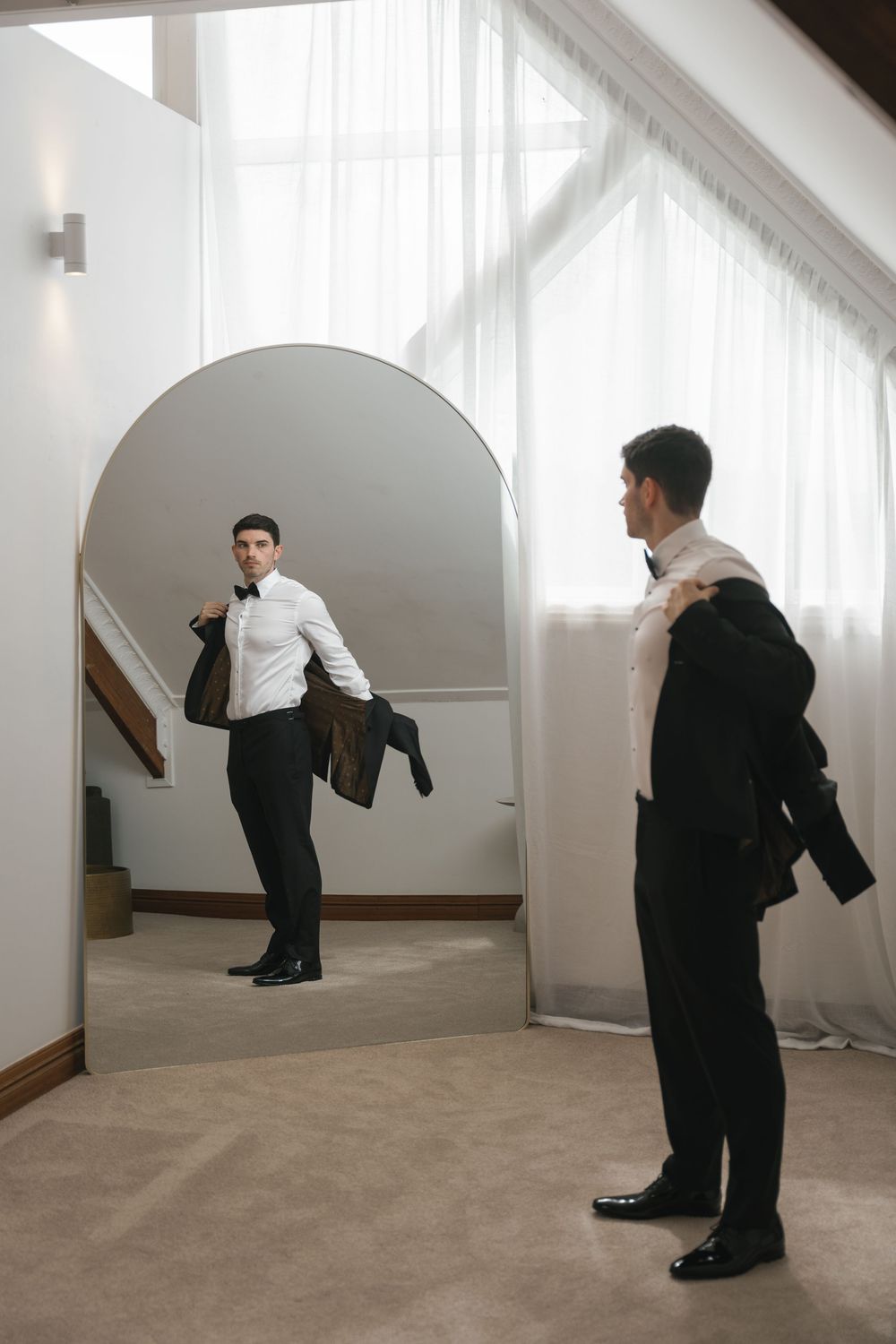 A groom in a white shirt and black suit checks his appearance in a large arched mirror while holding his jacket.
