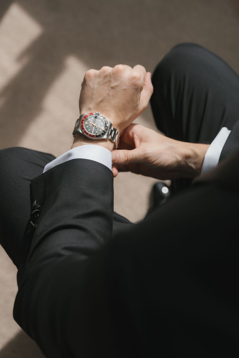 Man in black suit checking luxury analog wristwatch with red and silver bezel, close-up detail shot.